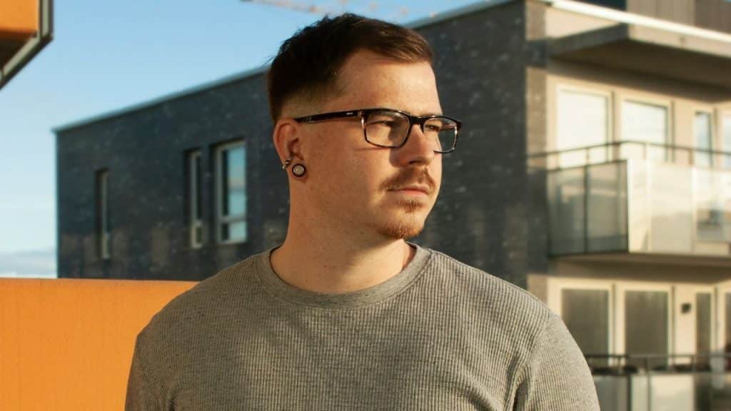 Man wearing glasses and gauges, looking away, with modern apartment buildings in the background.