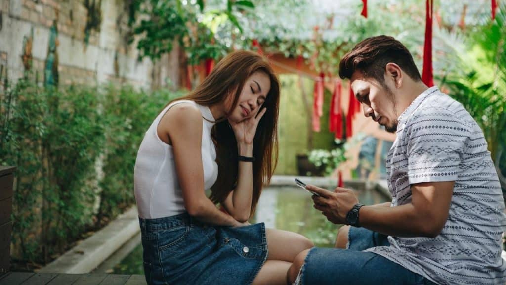 Woman with eyes closed resting her head on her hand next to a man looking at a phone outdoors.