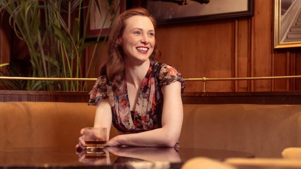 Smiling woman in a floral dress sitting at a table with a drink in a dimly lit, wood-paneled booth.