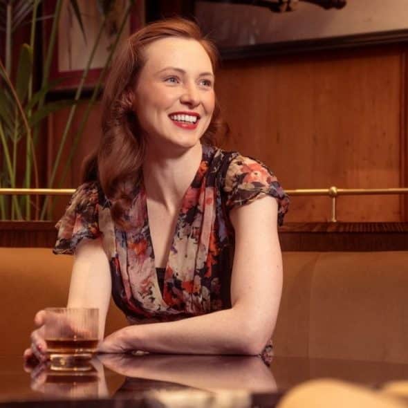 Smiling woman in a floral dress sitting at a table with a drink in a dimly lit, wood-paneled booth.