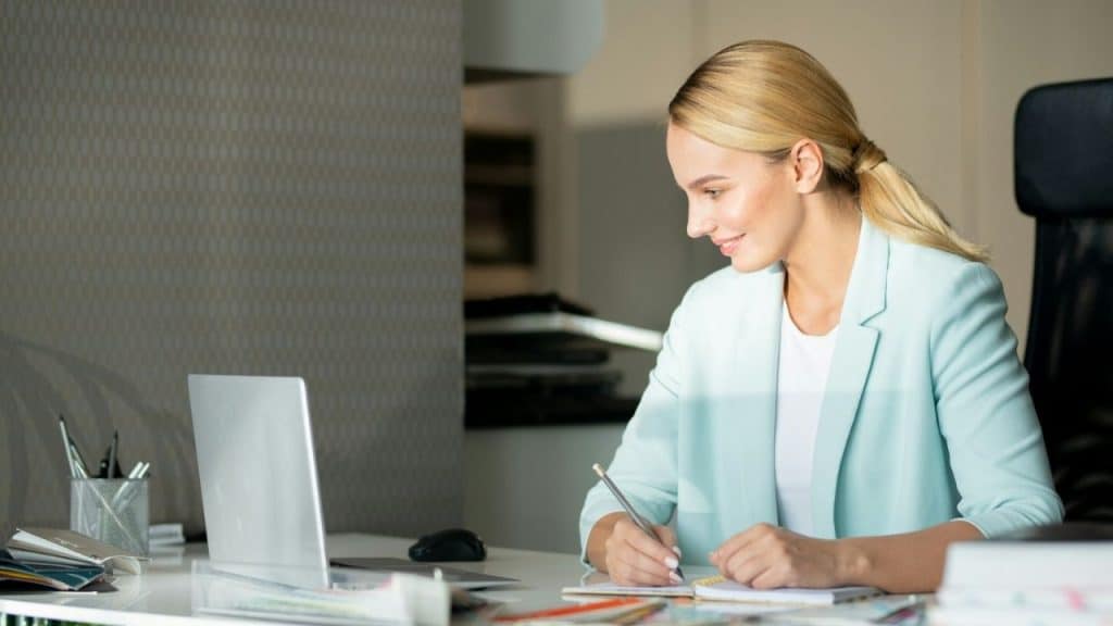 Blonde woman in a light blue blazer writing in a notebook while working on a laptop at a desk.