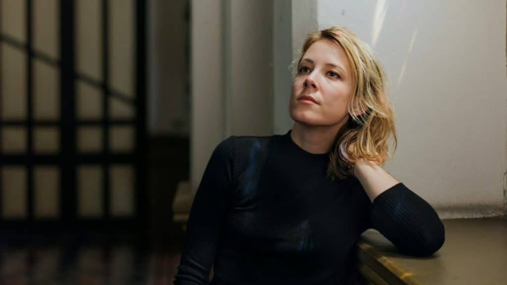 Blonde woman in a black top looking up thoughtfully in a dimly lit indoor space.
