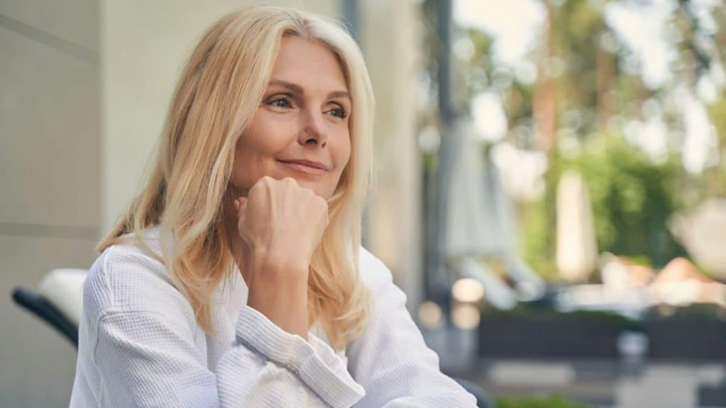 Middle-aged blonde woman in a white robe smiling and looking away, with her chin resting on her fist.