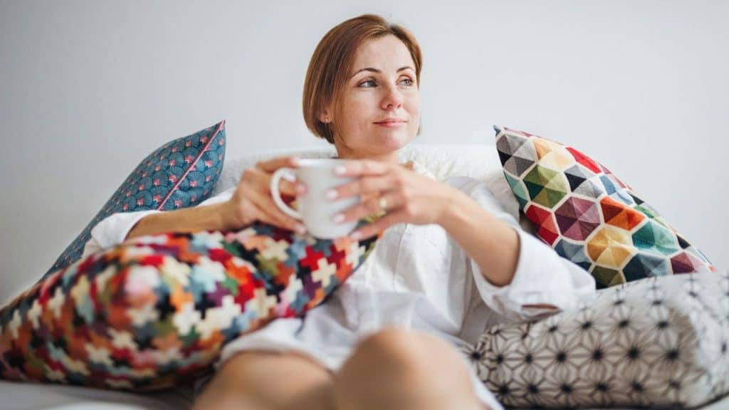 Woman relaxing on a couch surrounded by patterned pillows, holding a white mug.