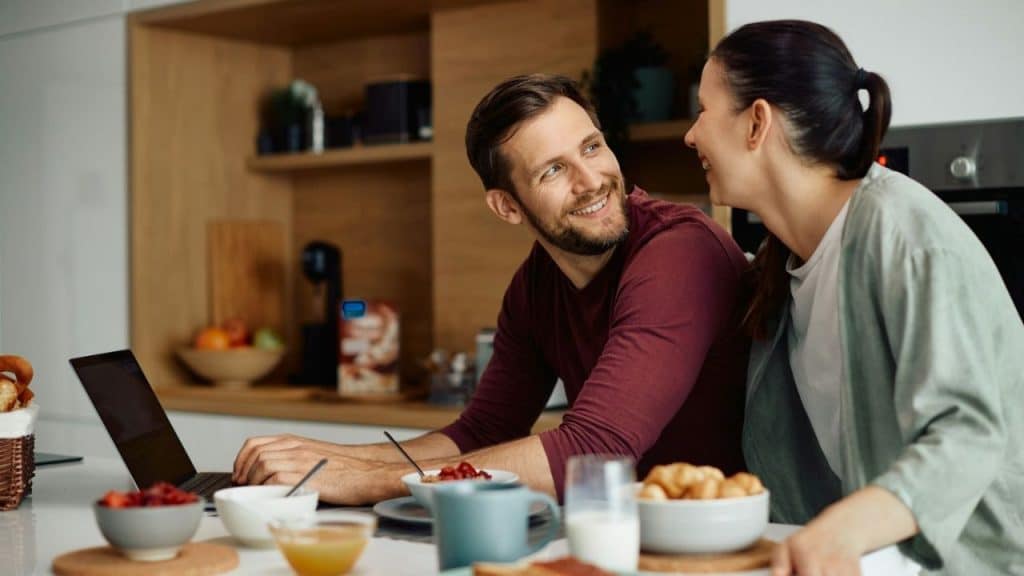 Happy couple smiling at each other over a laptop and breakfast in a modern kitchen.