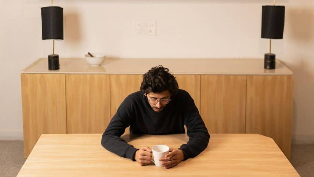 A man with curly hair sits alone at a wooden table, holding a mug and looking down.