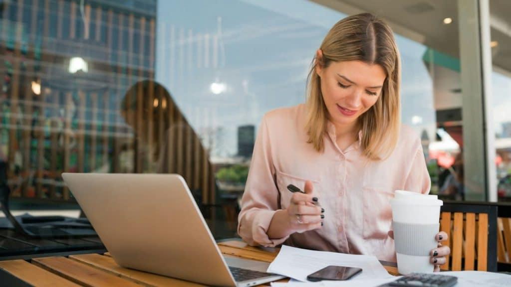 Woman reviewing documents with a pen and coffee cup, sitting with a laptop outdoors.