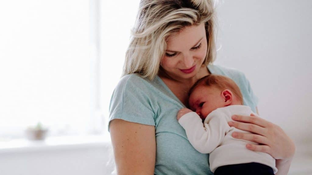 Blonde woman in a light blue shirt holding a red-haired baby in a white sleeper against her chest.