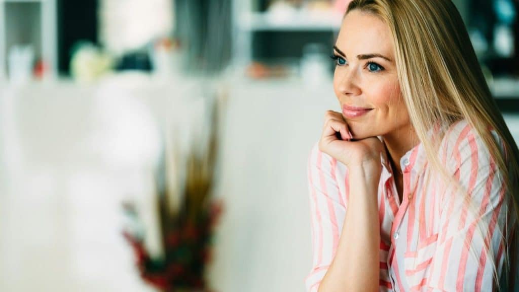Blonde woman with blue eyes smiling, resting her chin on her hand, in a soft-focus indoor setting.