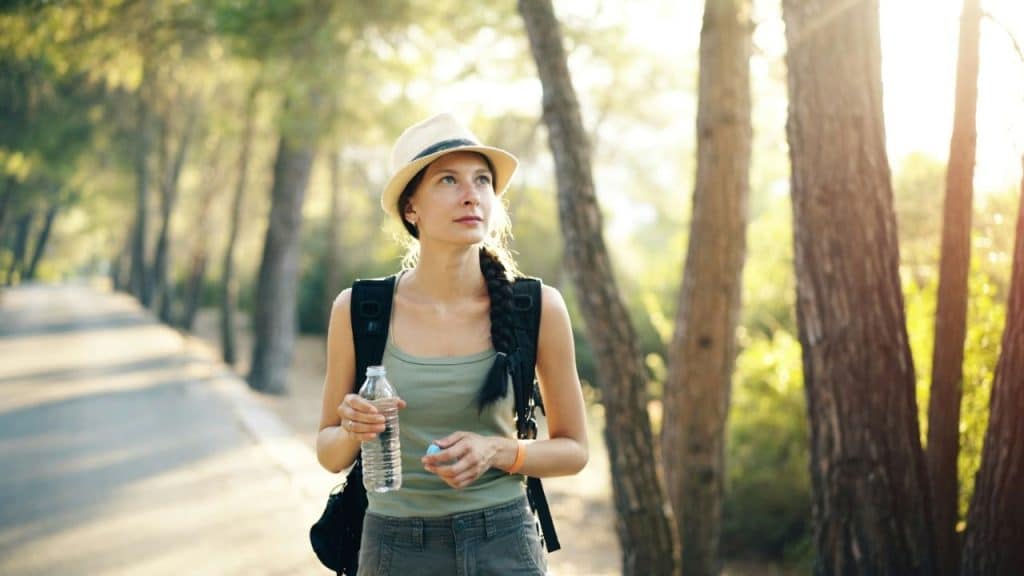 A woman with a backpack and sun hat hiking on a tree-lined path, holding a water bottle.