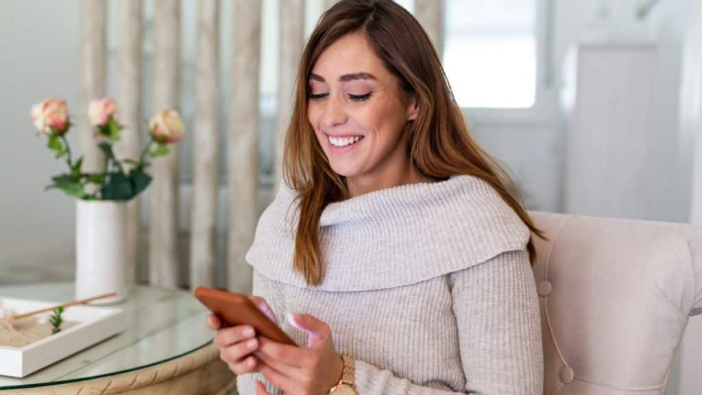 Smiling woman in a light sweater looking at her phone while sitting indoors.