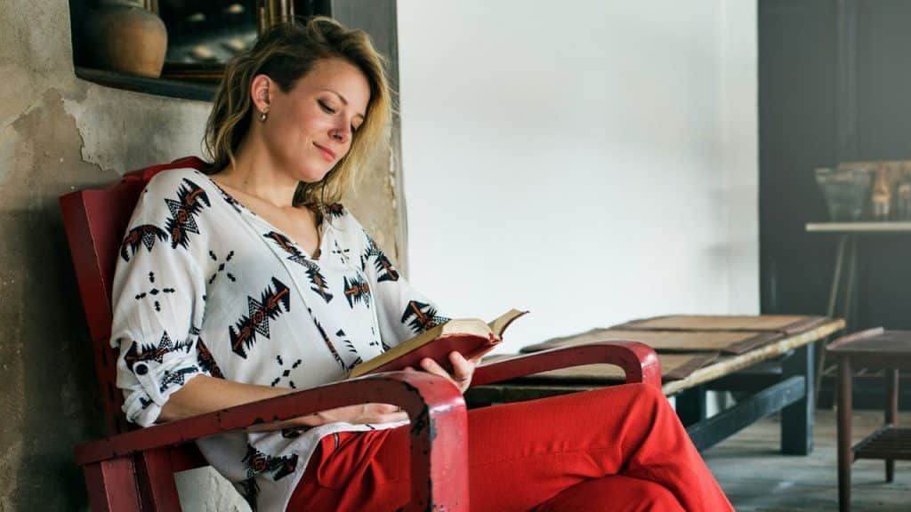 Young woman in a patterned white top and red pants sitting in a red chair reading a book indoors.