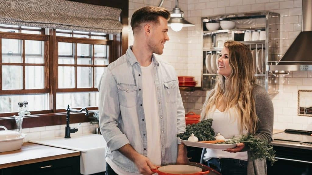 A smiling man and woman standing in a kitchen, holding cooking items and looking at each other.