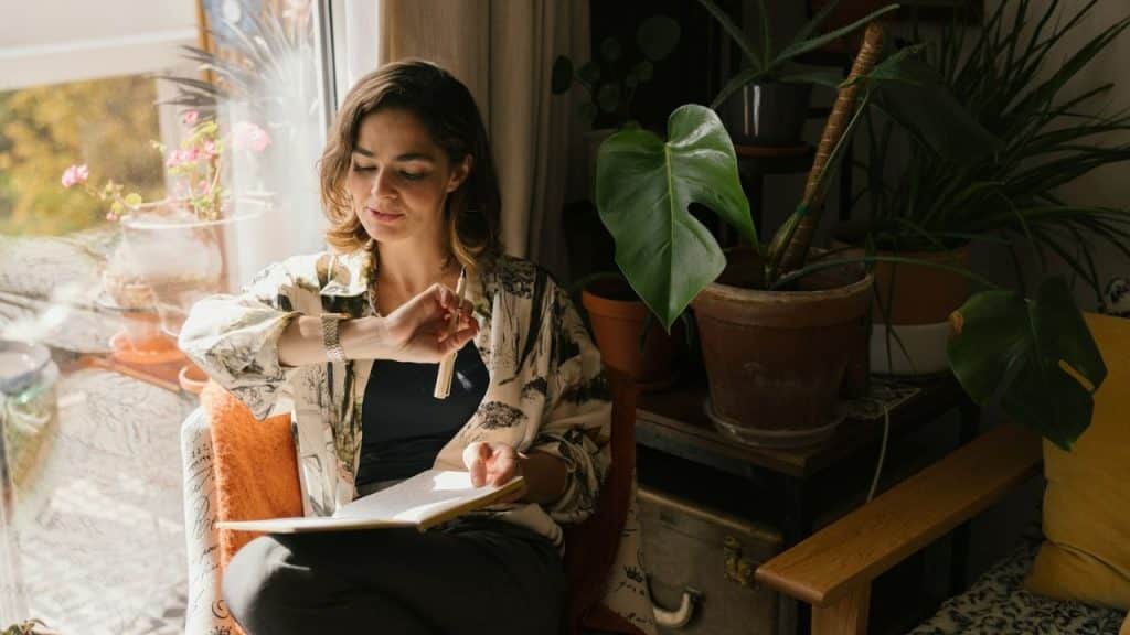 Woman looking at her watch while writing in a notebook, sitting by a window surrounded by houseplants.