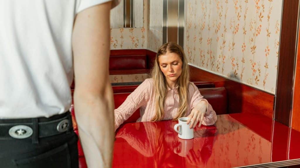 Woman seated in a retro diner booth with a red table, looking down at a coffee mug, as a server stands near her.