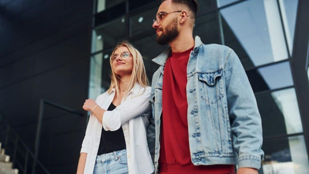 A casually dressed couple with glasses stands close together outside a modern building.
