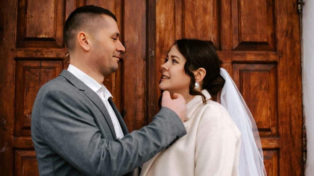 A newly married couple, the groom in a gray suit and the bride in a white coat and veil, smiling at each other.