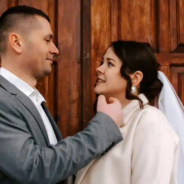 A newly married couple, the groom in a gray suit and the bride in a white coat and veil, smiling at each other.