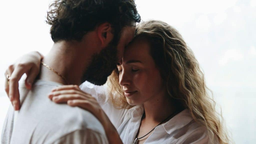 A bearded man and a woman with long wavy hair embracing, forehead to forehead, with their eyes closed.