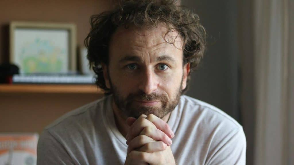 Man with curly hair and a beard, in a light shirt, looking directly at the camera with clasped hands.