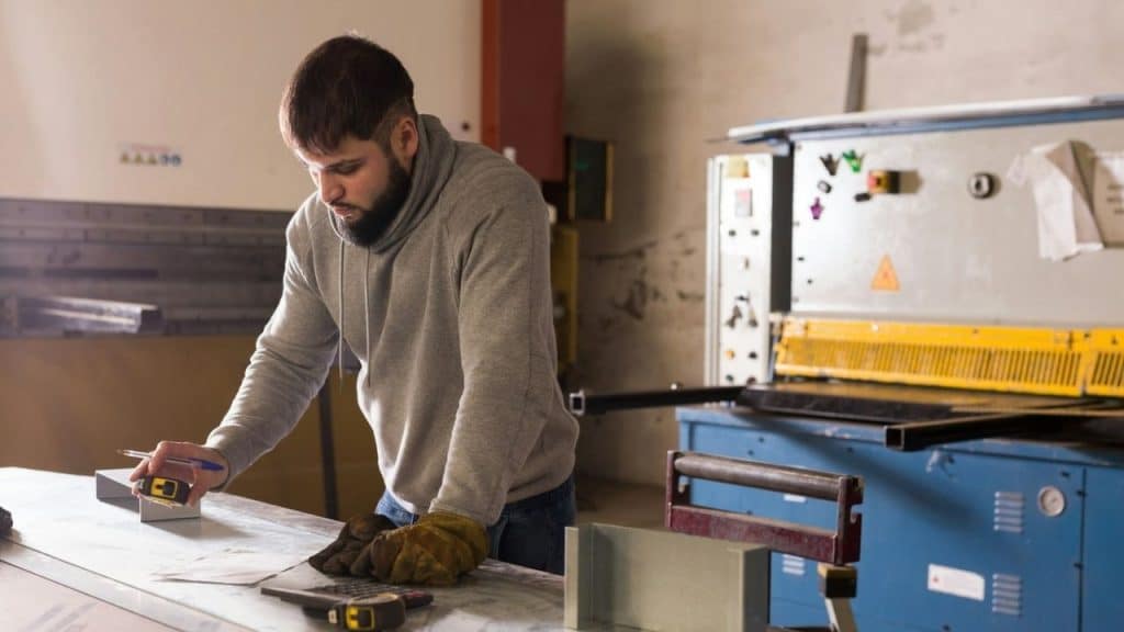 Bearded man in a gray hoodie measuring material on a workbench next to a large blue and yellow machine in a workshop.