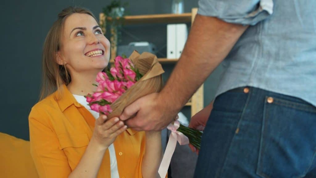 Woman smiling excitedly while receiving a bouquet of pink flowers wrapped in brown paper from a man.