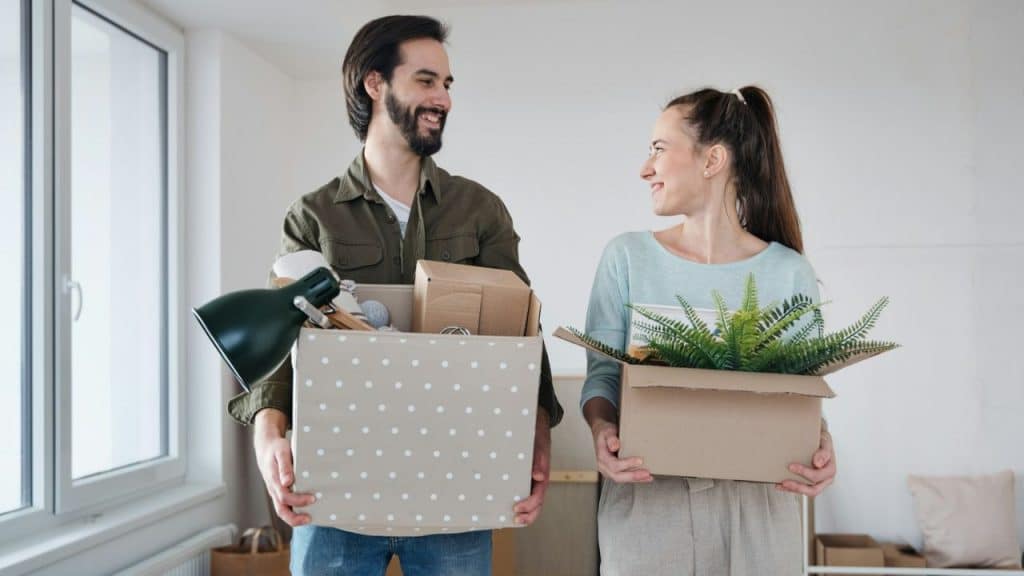 Smiling man and woman standing and carrying boxes with household items, looking at each other.