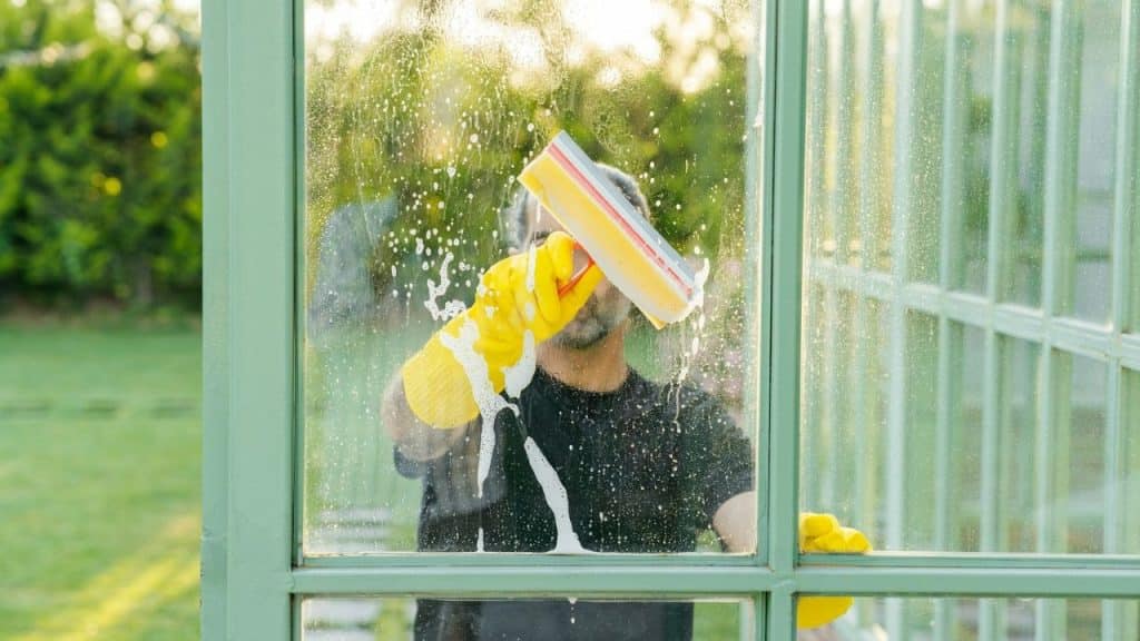 A person in yellow gloves using a yellow squeegee to clean a window of a green structure outdoors.