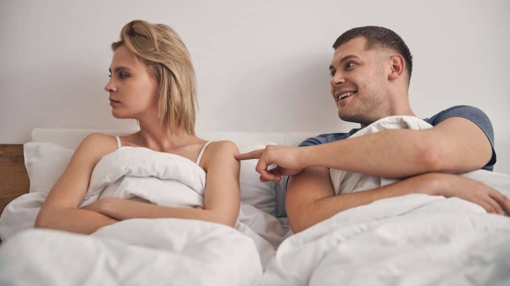 A woman with her back turned ignores a smiling man who points at her shoulder in bed.