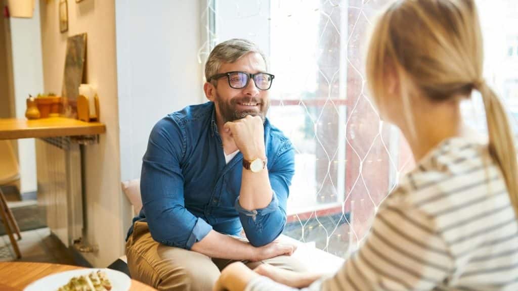 Man with a beard and glasses smiling while sitting at a table and talking to a person with blonde hair.