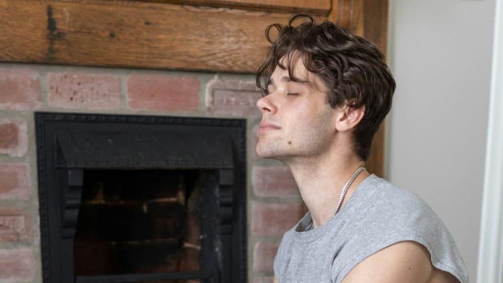 Young man with brown, wavy hair, in a gray t-shirt, sitting with his eyes closed near a brick fireplace.