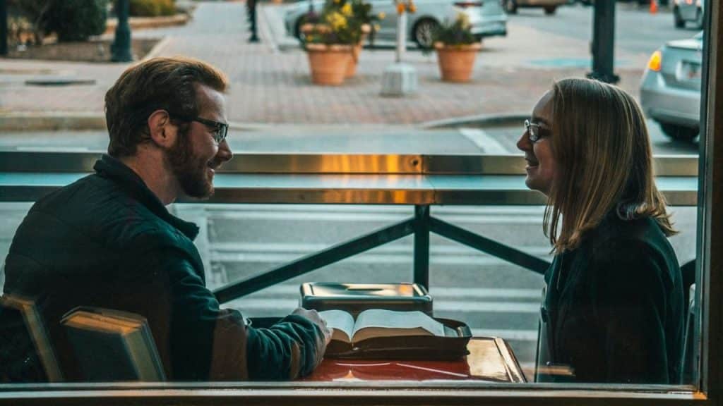 A man and woman in glasses smiling at each other across a table with an open book, viewed through a window.