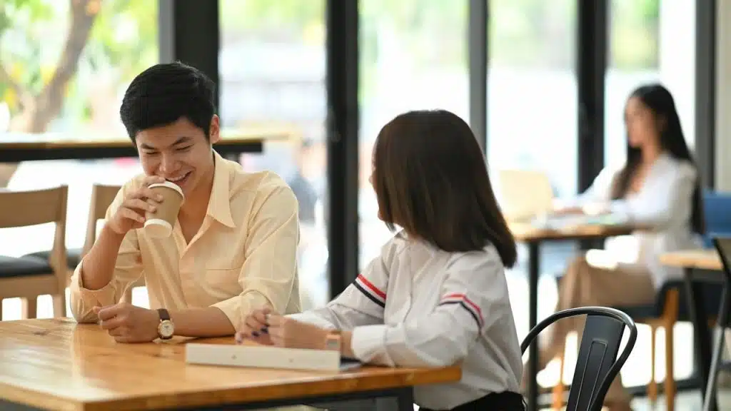 A man drinking coffee and smiling while sitting at a table in a cafe with a woman.