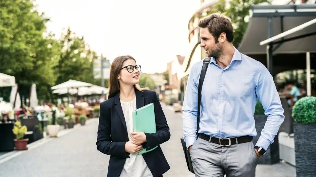 A woman with glasses and holding a file walking down a street with a man with a shoulder bag.