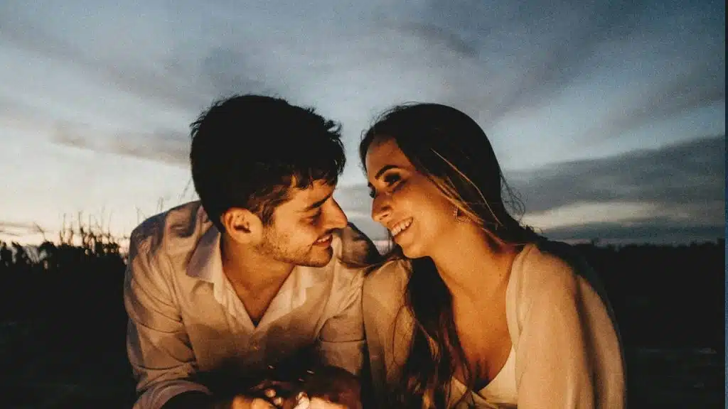 A man looking at a woman while their faces are illuminated by the light from a fire at dusk.