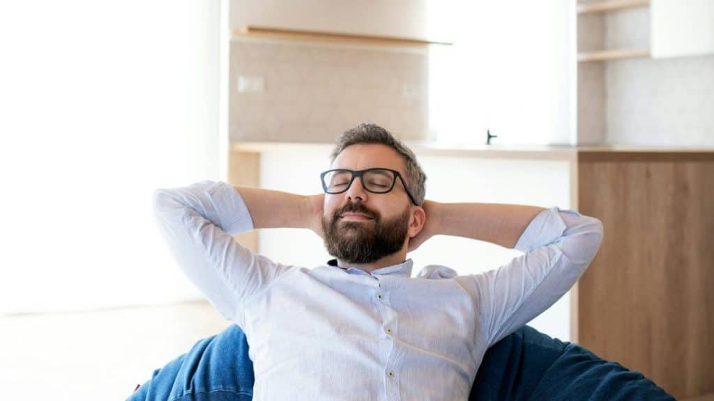 A relaxed, bearded man with glasses leans back with his hands behind his head.