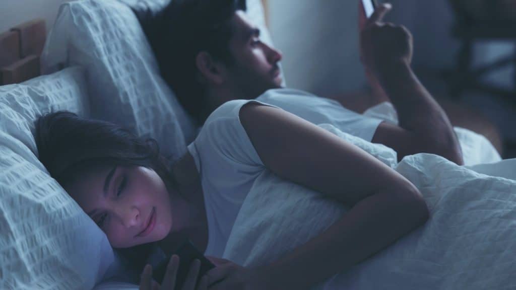 A man and woman lie in bed in a dark room, both using their phones under the covers.