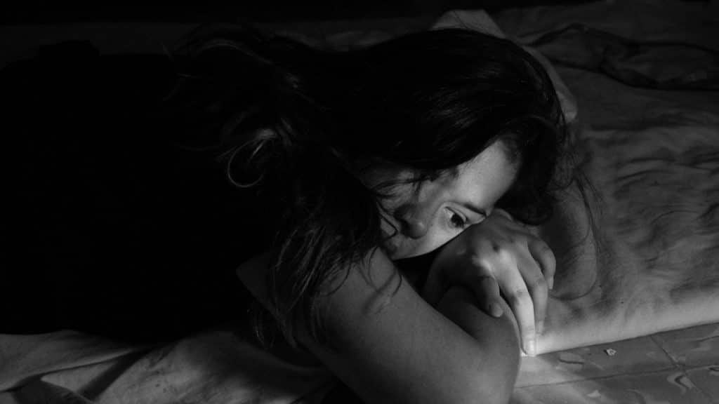 A high-contrast, black and white photo of a woman lying face-down on a bed, looking down sadly or pensively.