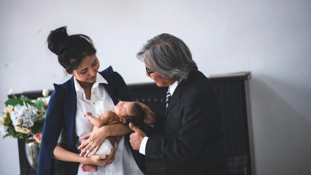 A professional-looking woman in a blazer is holding a newborn baby while a smiling older man in a suit and tie looks on and gently touches the baby's head.