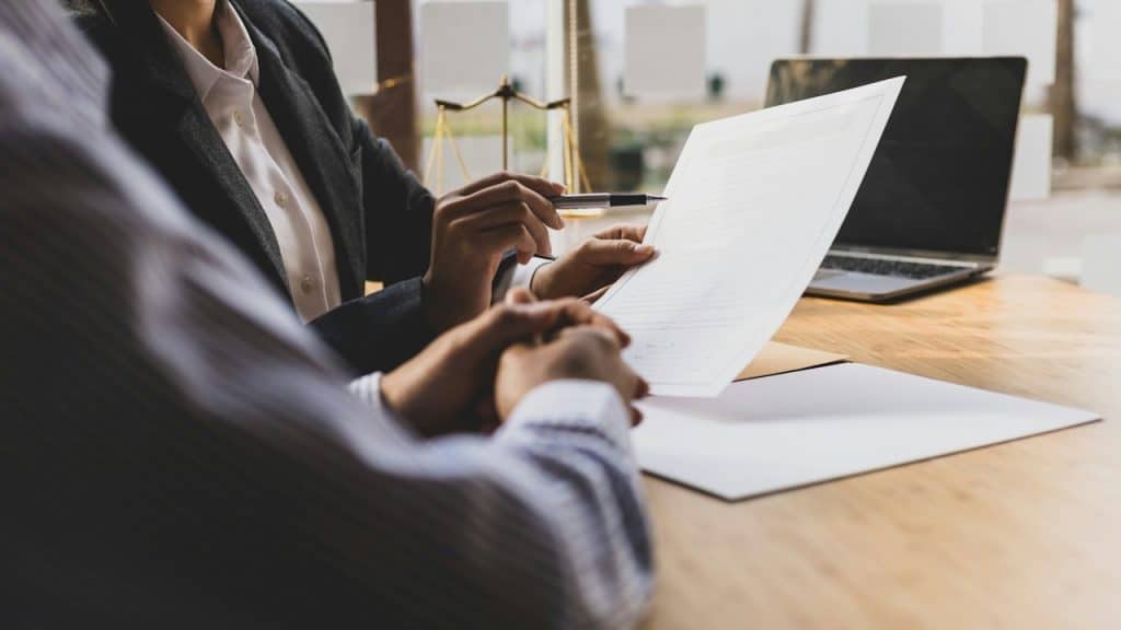A lawyer or financial advisor in a suit is pointing with a pen to a document, explaining a contract to a client sitting across a wooden desk.