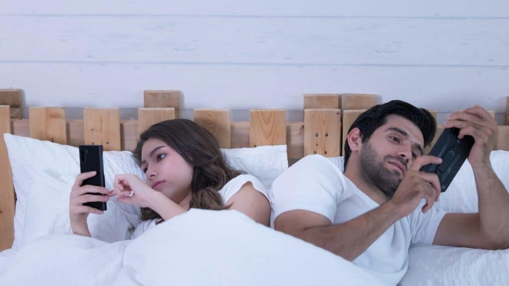 A man and a woman lie in bed back-to-back, both preoccupied with looking at their mobile phones.
