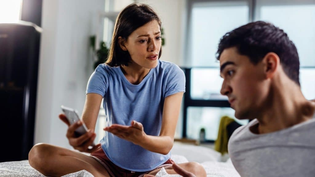 A stressed woman sits on a bed, holding a phone and gesturing angrily while arguing with a man sitting in the foreground.