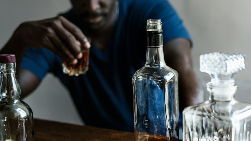 A blurry man in a blue shirt is seated, holding a shot glass, with several full and empty bottles of liquor in the foreground.