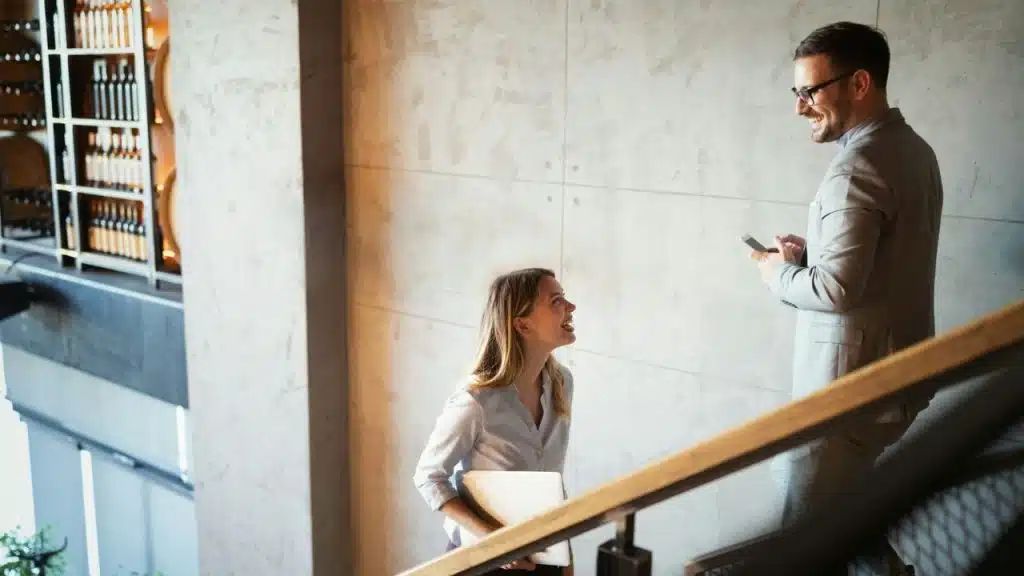 A man and a woman in business attire smile at each other while walking up a staircase.