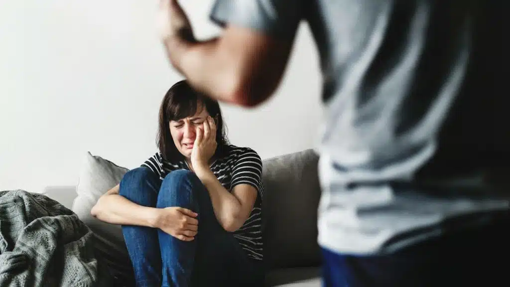 A distraught woman is crying on a couch with her hands over her face, while an out-of-focus man stands over her with an uplifted hand.