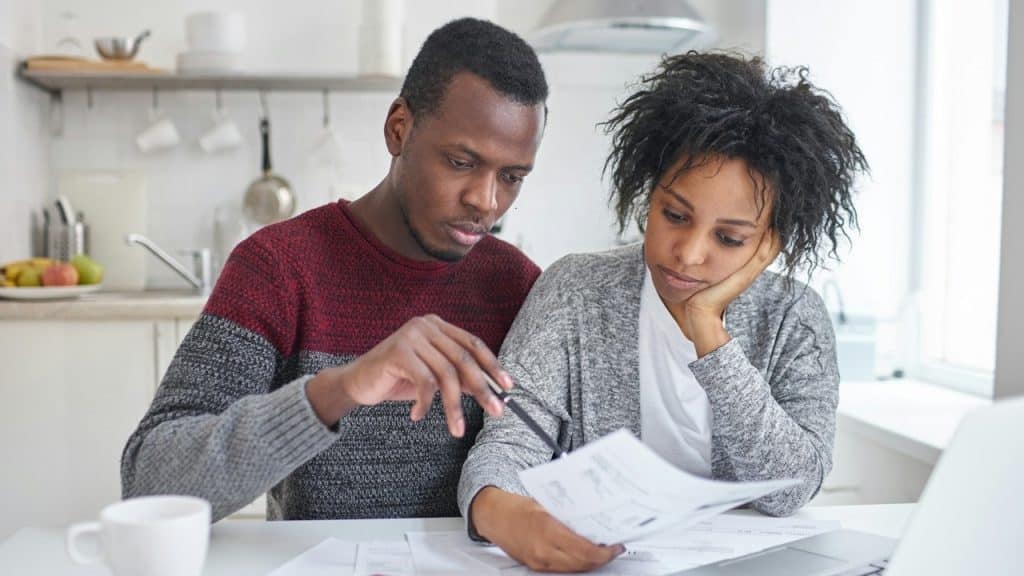 A focused couple sits at a white table in a kitchen, looking stressed as they review paperwork and bills with a laptop and calculator nearby.