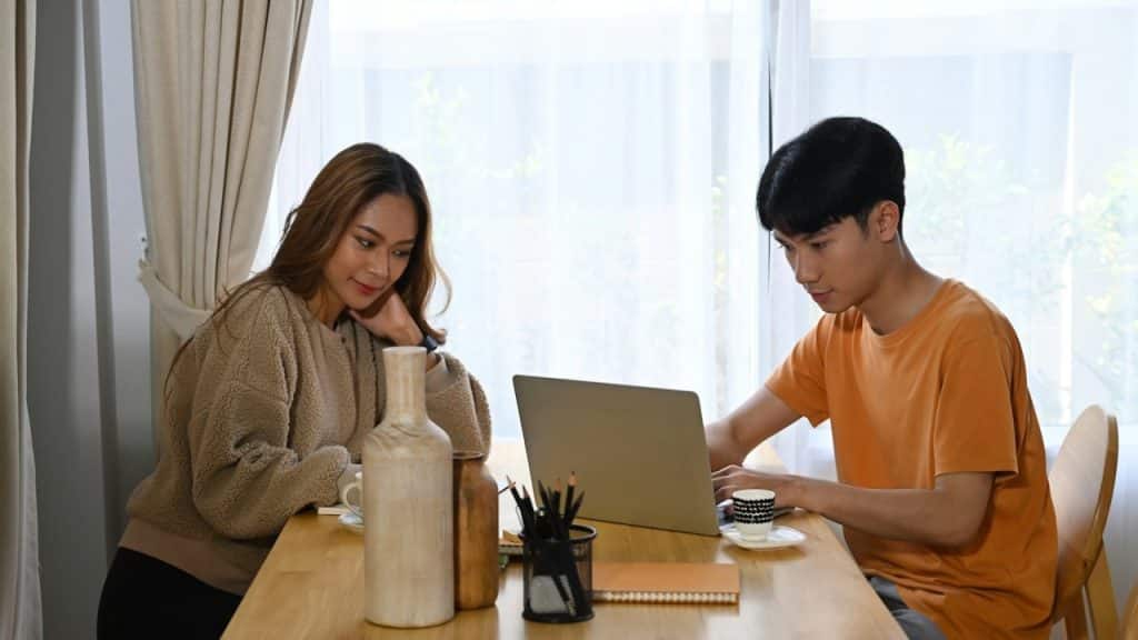 A young man and woman are sitting at a wooden table in a bright room, looking at a laptop together.