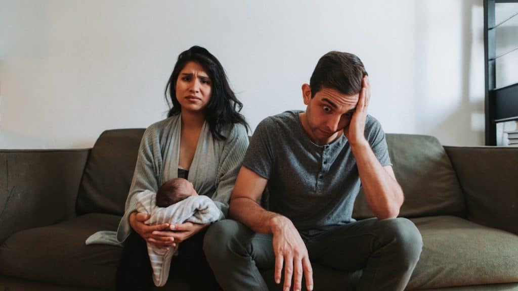 A young, stressed-looking couple sits on a couch, with the woman holding a newborn baby while the man holds his head in his hand