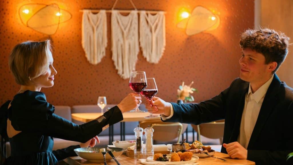 A man and woman dressed up for a date night are clinking glasses of red wine over a table with food in a warmly lit restaurant.