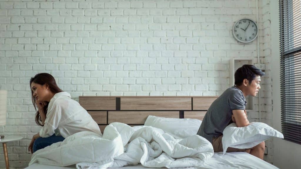 A man and a woman sit on opposite sides of a bed, both looking away from each other with pensive or upset expressions